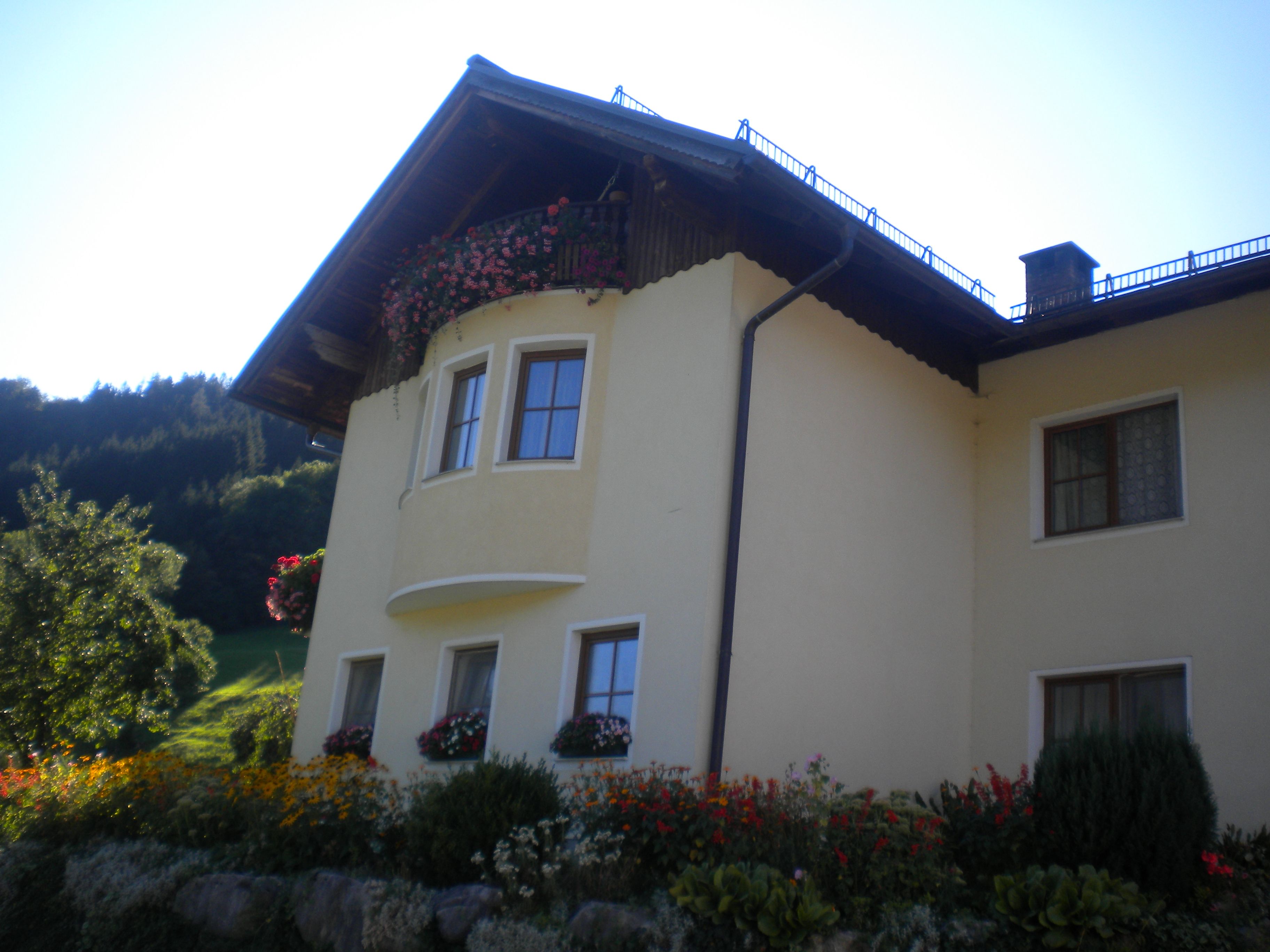 Gelbes Haus mit Blumen und Balkon vor bewaldetem Hügel.