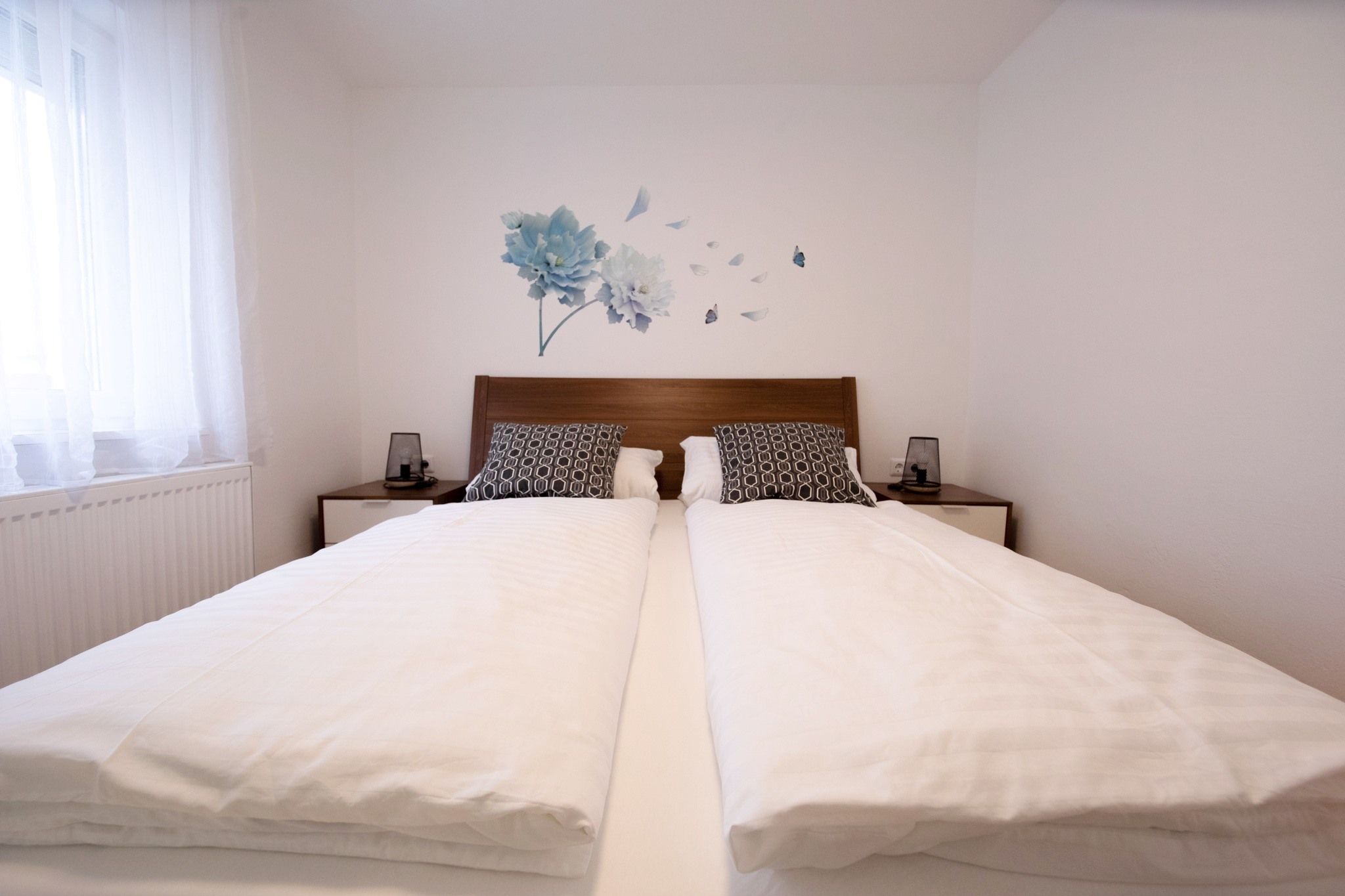 Ein modernes Schlafzimmer mit einem Doppelbett, weißen Bettdecken und dekorativen Kissen. An der Wand ist ein Blumenmotiv zu sehen.