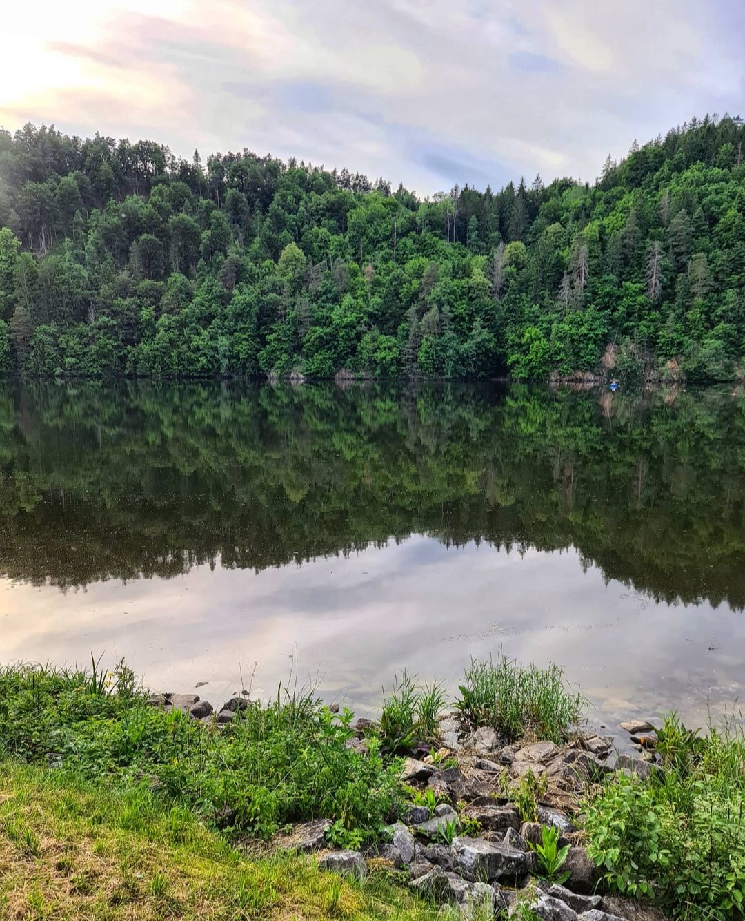 Ein ruhiger See mit bewaldeten Hügeln im Hintergrund, die sich im Wasser spiegeln.