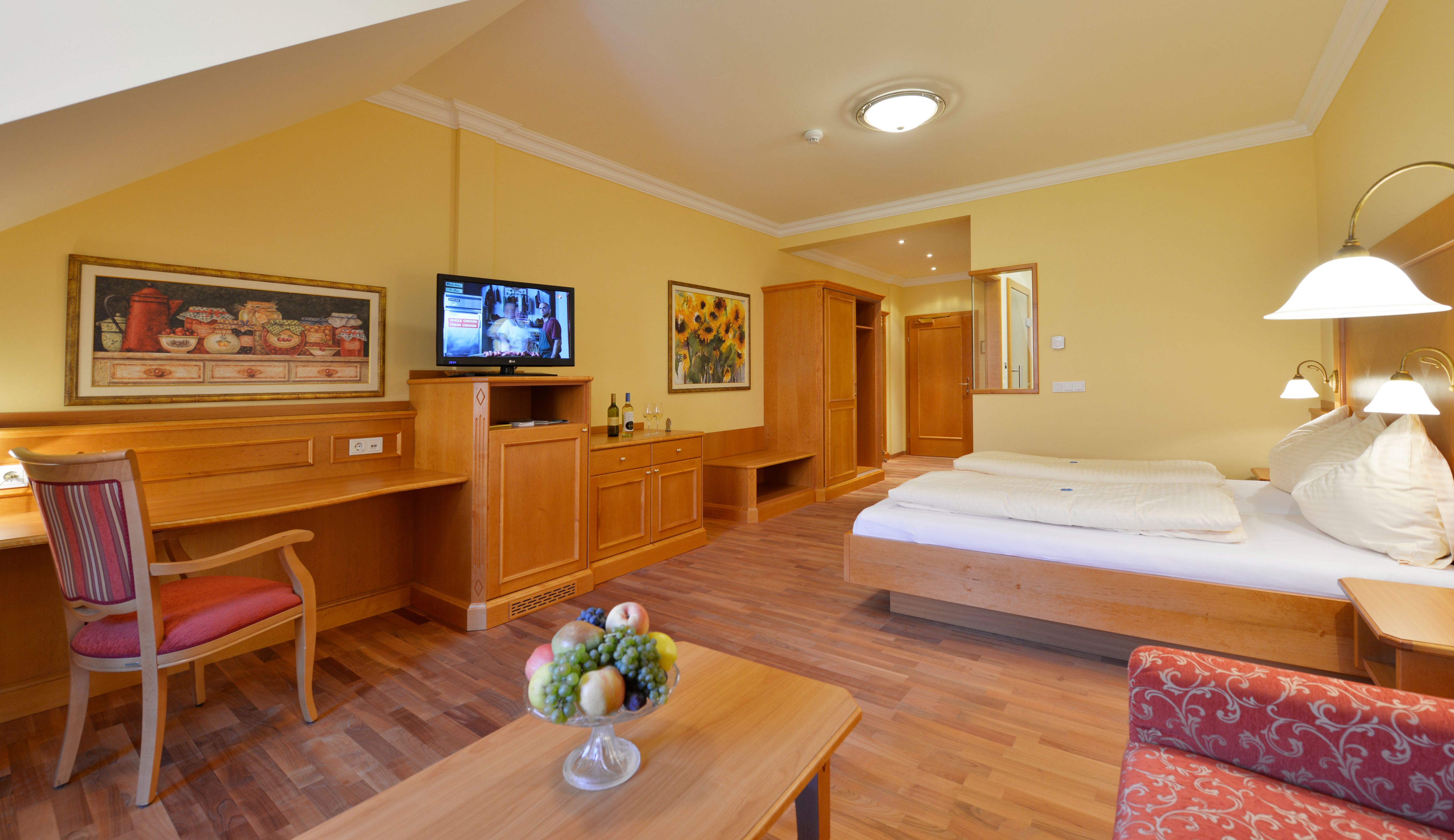Ein gemütliches Gästezimmer mit Holzmöbeln, einem Doppelbett, einem Schreibtisch, einem Fernseher und einem Obstteller auf dem Tisch.