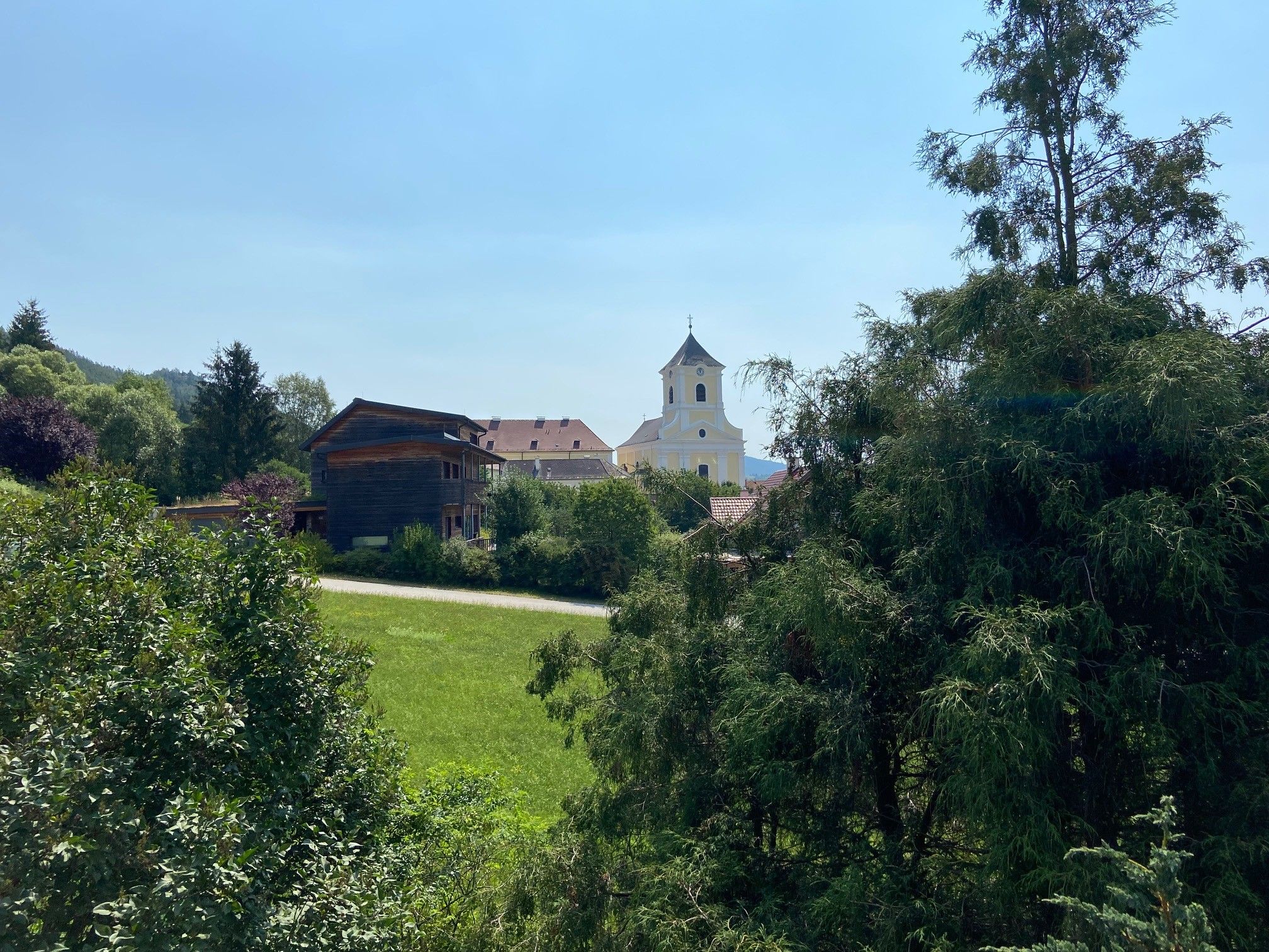 Blick auf eine Kirche und ein Holzhaus hinter Bäumen und Wiesen.