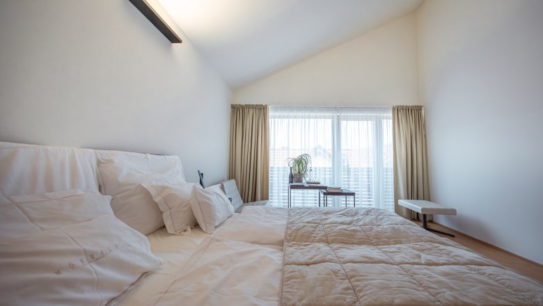 Modernes Hotelzimmer mit gro&szlig;em Bett, wei&szlig;en Kissen und Tagesdecke, helle Vorh&auml;nge und Fenster im Hintergrund.