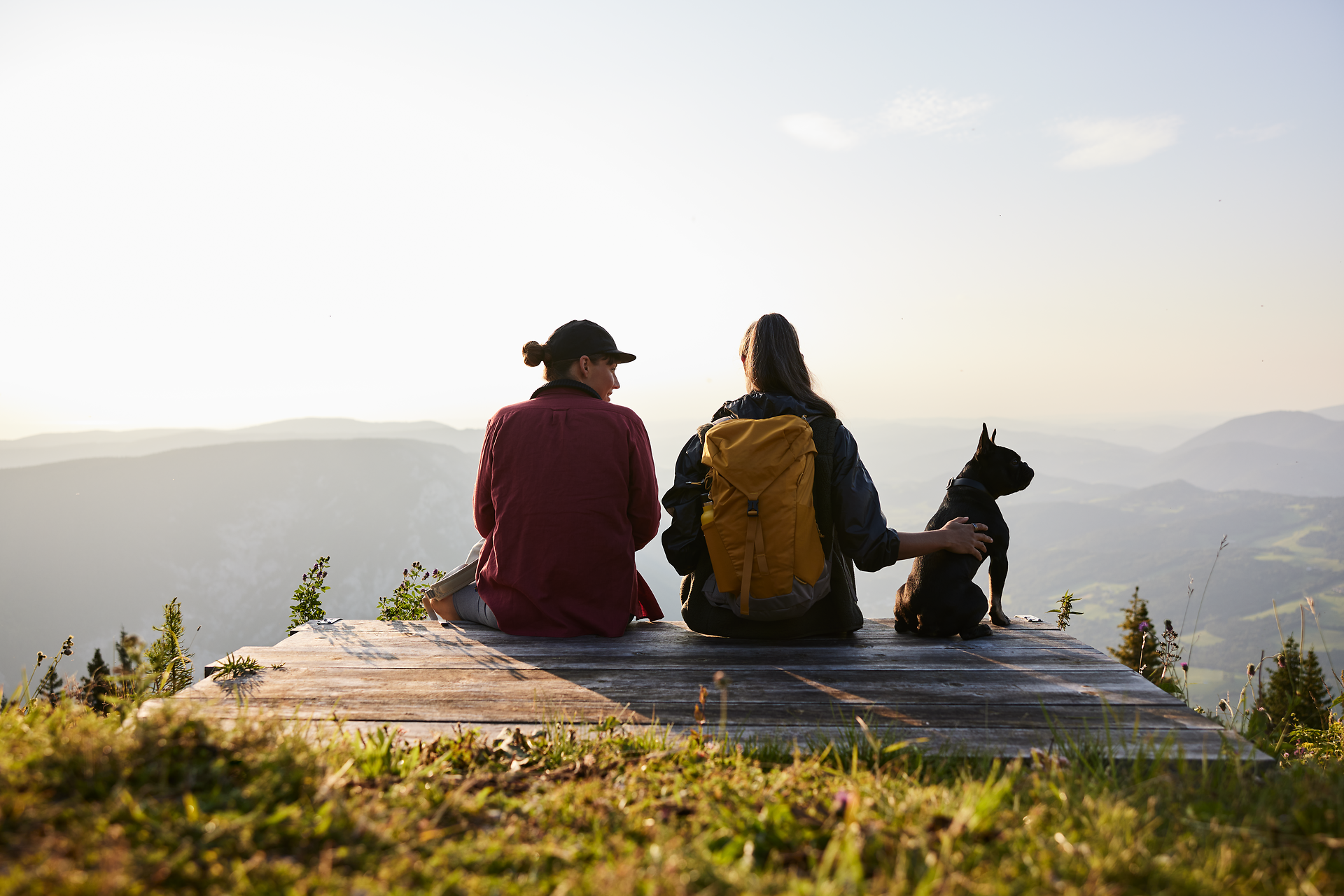In der goldenen Abendsonne genießen zwei Wanderer die atemberaubende Aussicht auf die sanften Hügel und das weite Tal. Ihr treuer Begleiter, ein kleiner Hund, sitzt entspannt neben ihnen und teilt diesen friedlichen Moment der Naturverbundenheit. Die frische Bergluft und die Stille der Umgebung laden dazu ein, die Seele baumeln zu lassen.