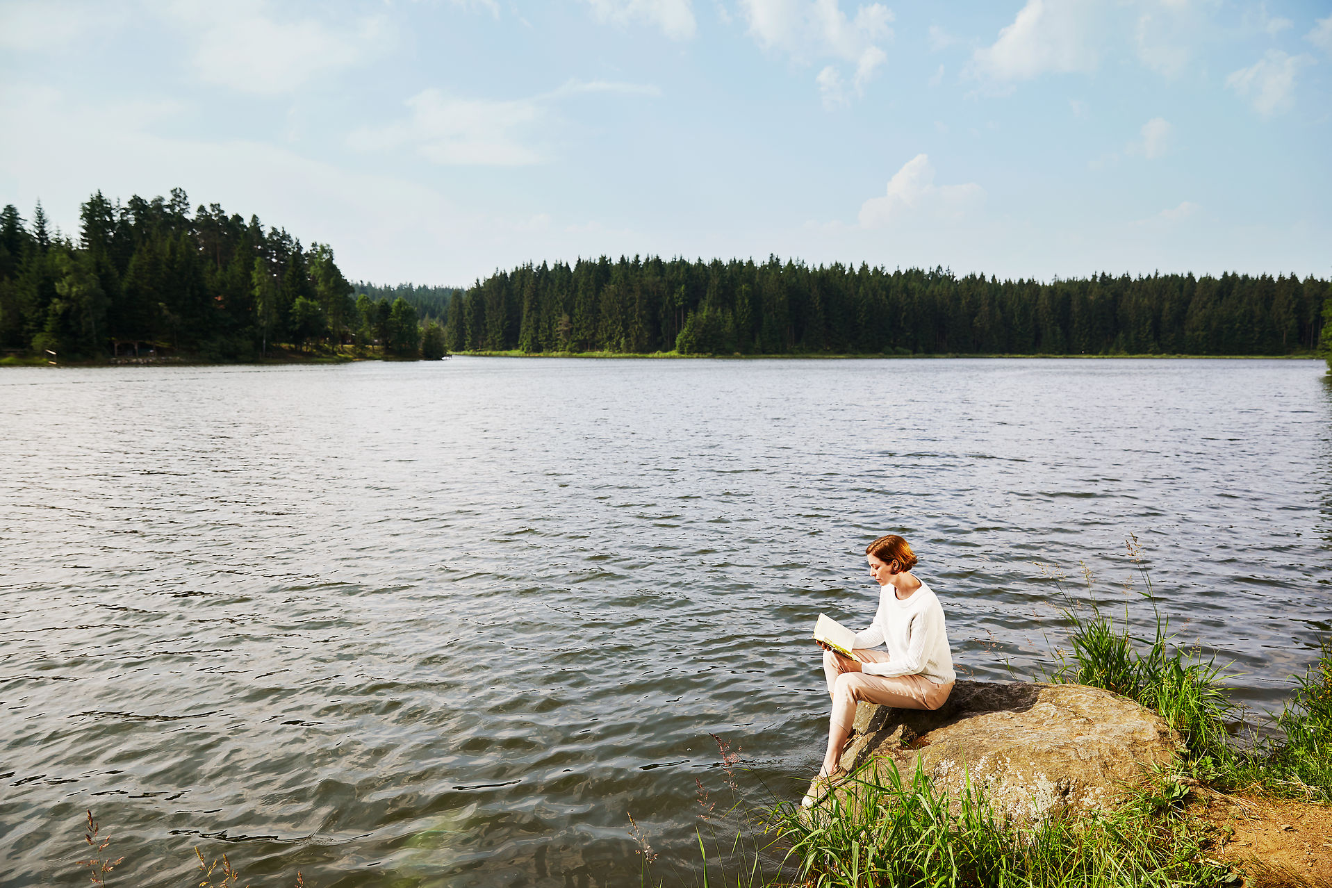 Am Ufer eines ruhigen Sees sitzt eine Person entspannt auf einem großen Stein und vertieft sich in ein Buch. Umgeben von üppigem Grün und dem sanften Plätschern des Wassers, strahlt die Szenerie eine friedliche Atmosphäre aus, die zum Verweilen einlädt.