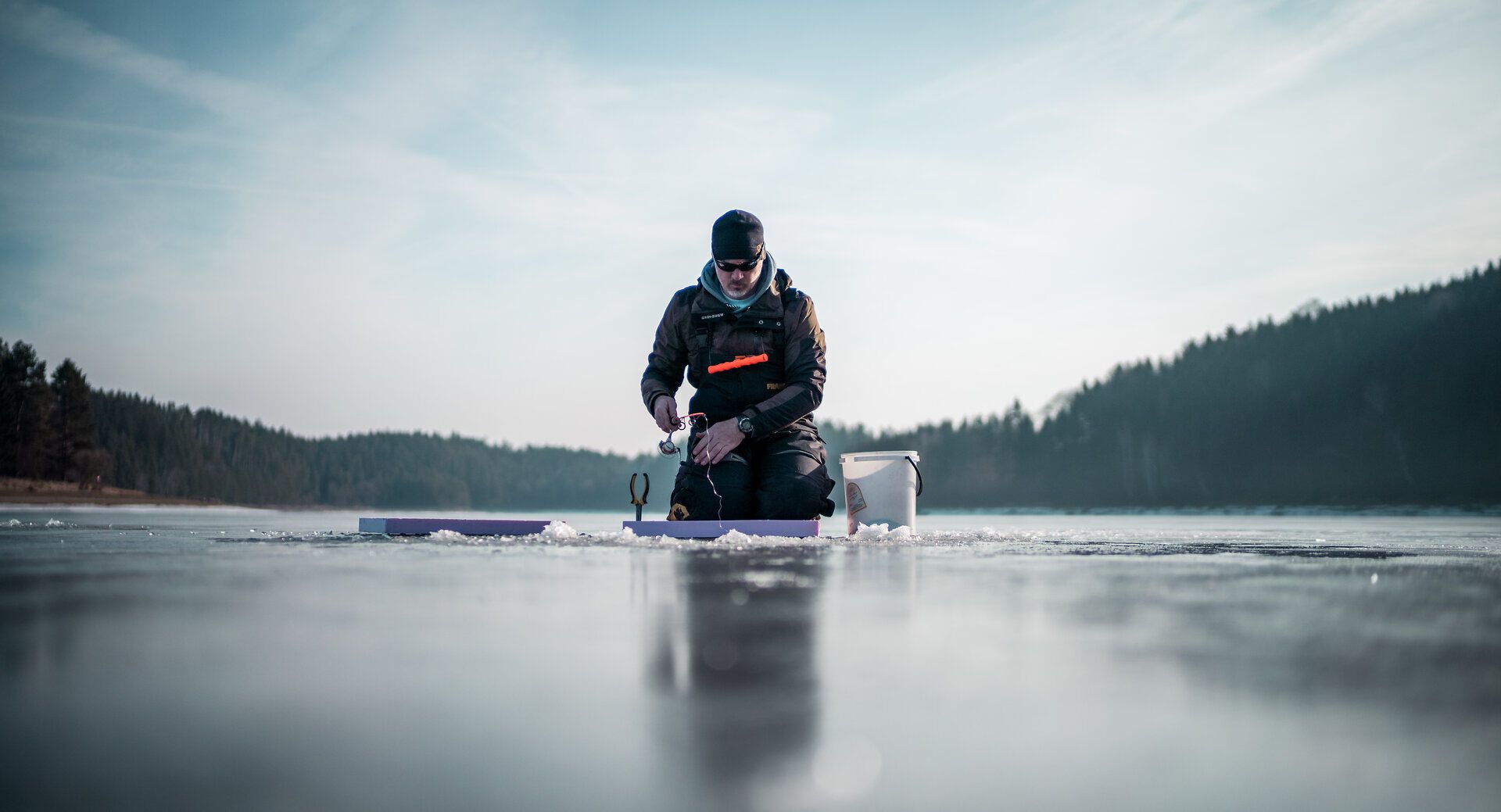 Ein Fischer sitzt konzentriert auf dem glitzernden Eis des Stausees Ottenstein und bereitet seine Angelausrüstung vor. Die ruhige Winterlandschaft, umgeben von schneebedeckten Bäumen, schafft eine friedliche Atmosphäre, die zum Entspannen einlädt. Hier, in der Stille der Natur, wird das Eisfischen zu einem unvergesslichen Erlebnis.