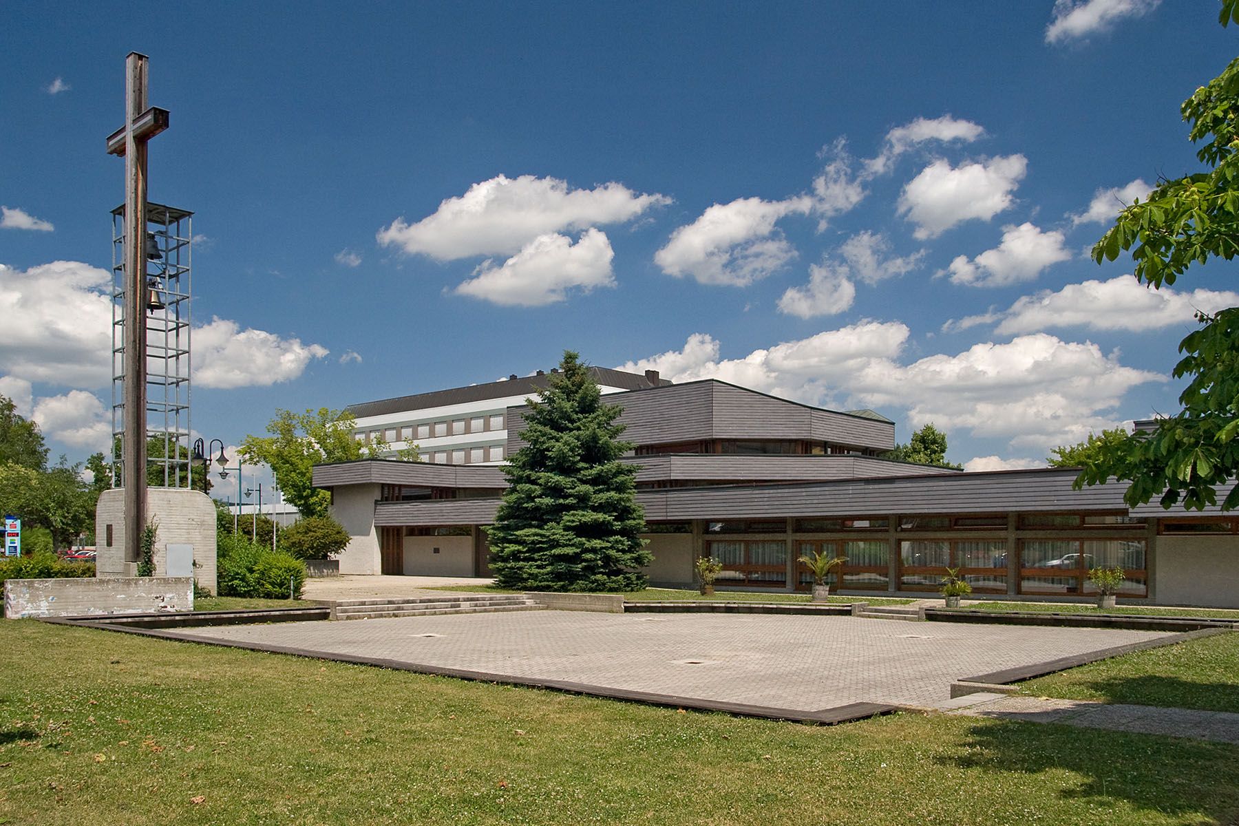 Moderne Kirche mit Glockenturm und Kreuz, umgeben von Grünflächen und Bäumen, unter blauem Himmel mit Wolken.