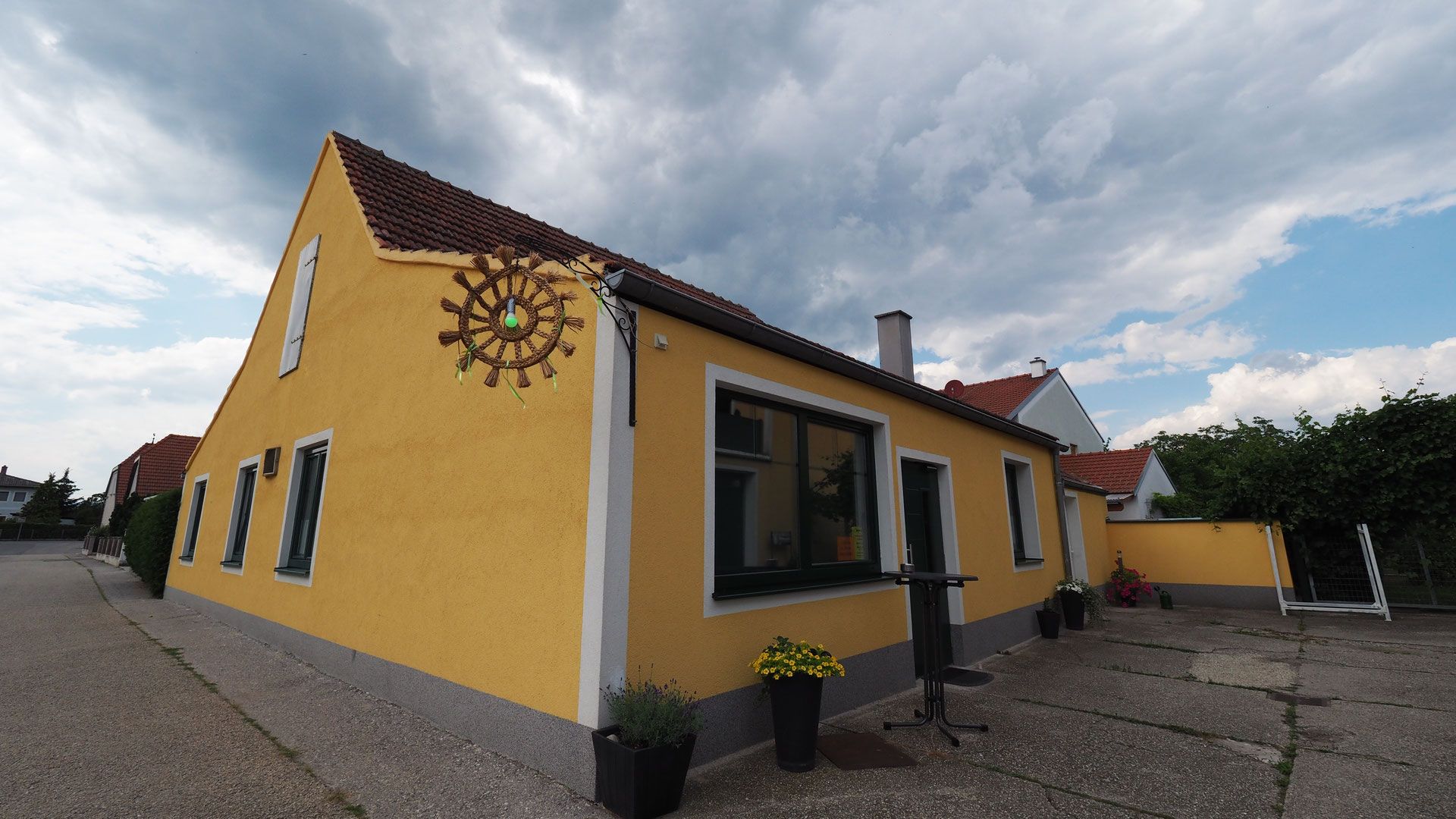 Gelbes Gebäude mit roten Dachziegeln und dekorativem Rad an der Wand, umgeben von Pflanzen und bewölktem Himmel.