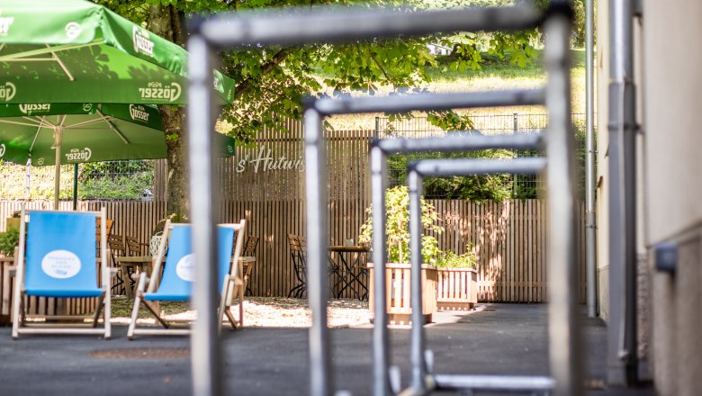 Blick auf einen Gastgarten mit grünen Sonnenschirmen und blauen Liegestühlen, umgeben von Bäumen und Holzzaun, im Vordergrund Radständer.