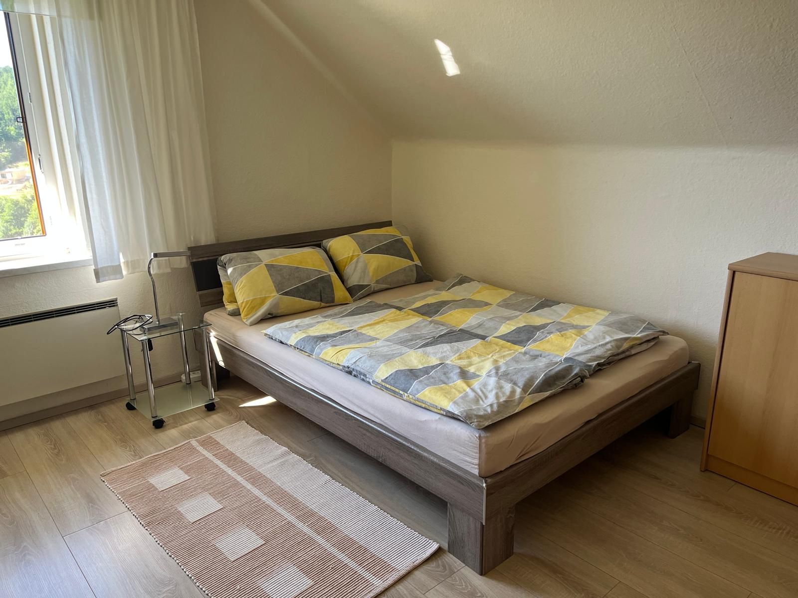 Ein gemütliches Schlafzimmer mit einem Doppelbett, gemusterten Bettbezügen, einem kleinen Nachttisch und einem Fenster mit Vorhängen.