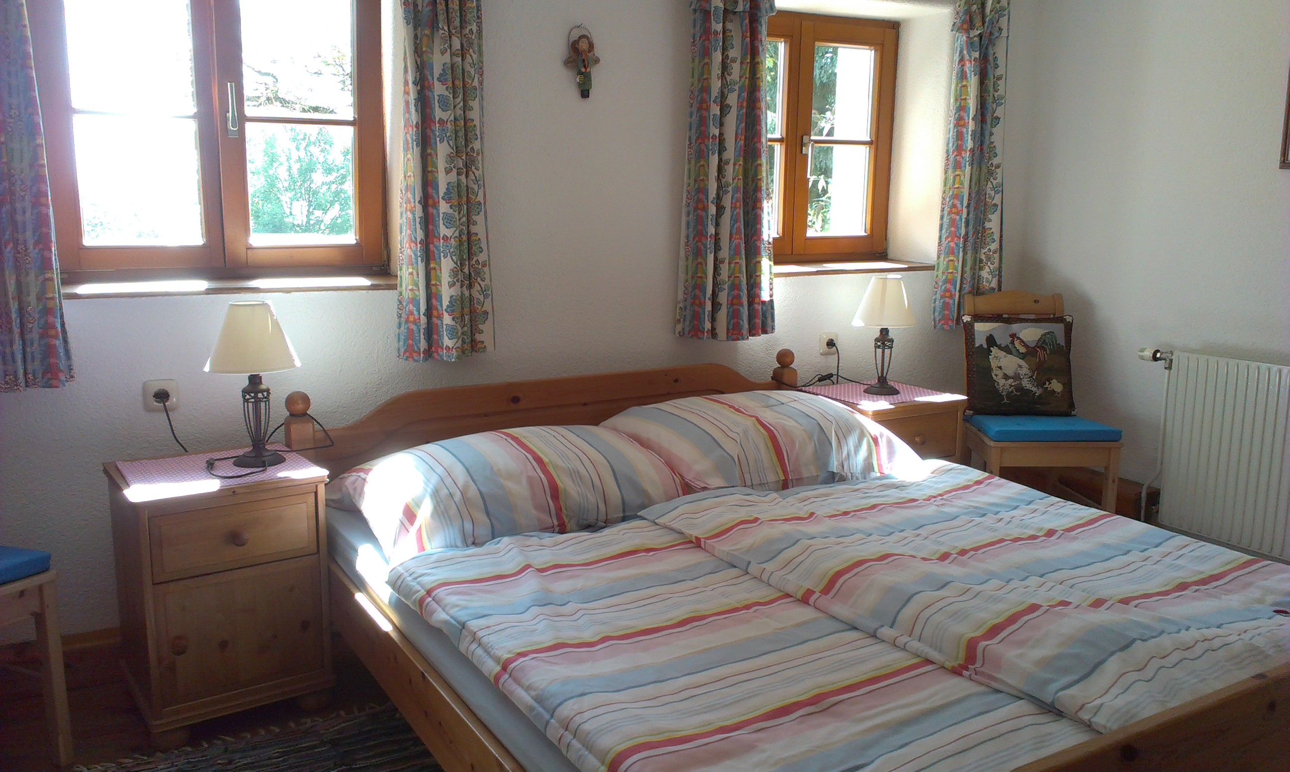 Gemütliches Schlafzimmer mit Doppelbett, gestreifter Bettwäsche, zwei Nachttischen mit Lampen und bunten Vorhängen an den Fenstern.