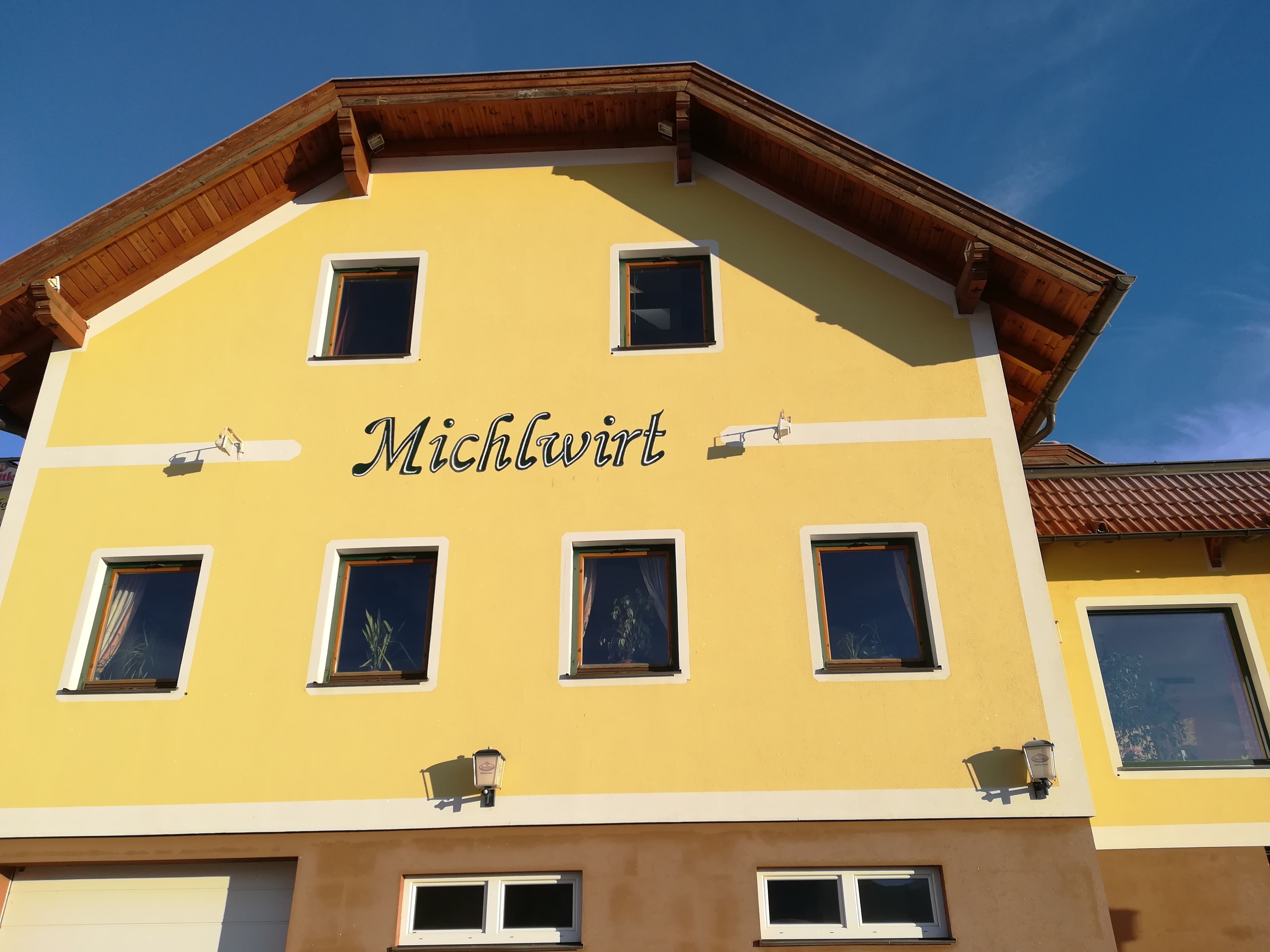 Gelbes Gebäude mit der Aufschrift 'Michlwirt' und mehreren Fenstern, unter blauem Himmel.