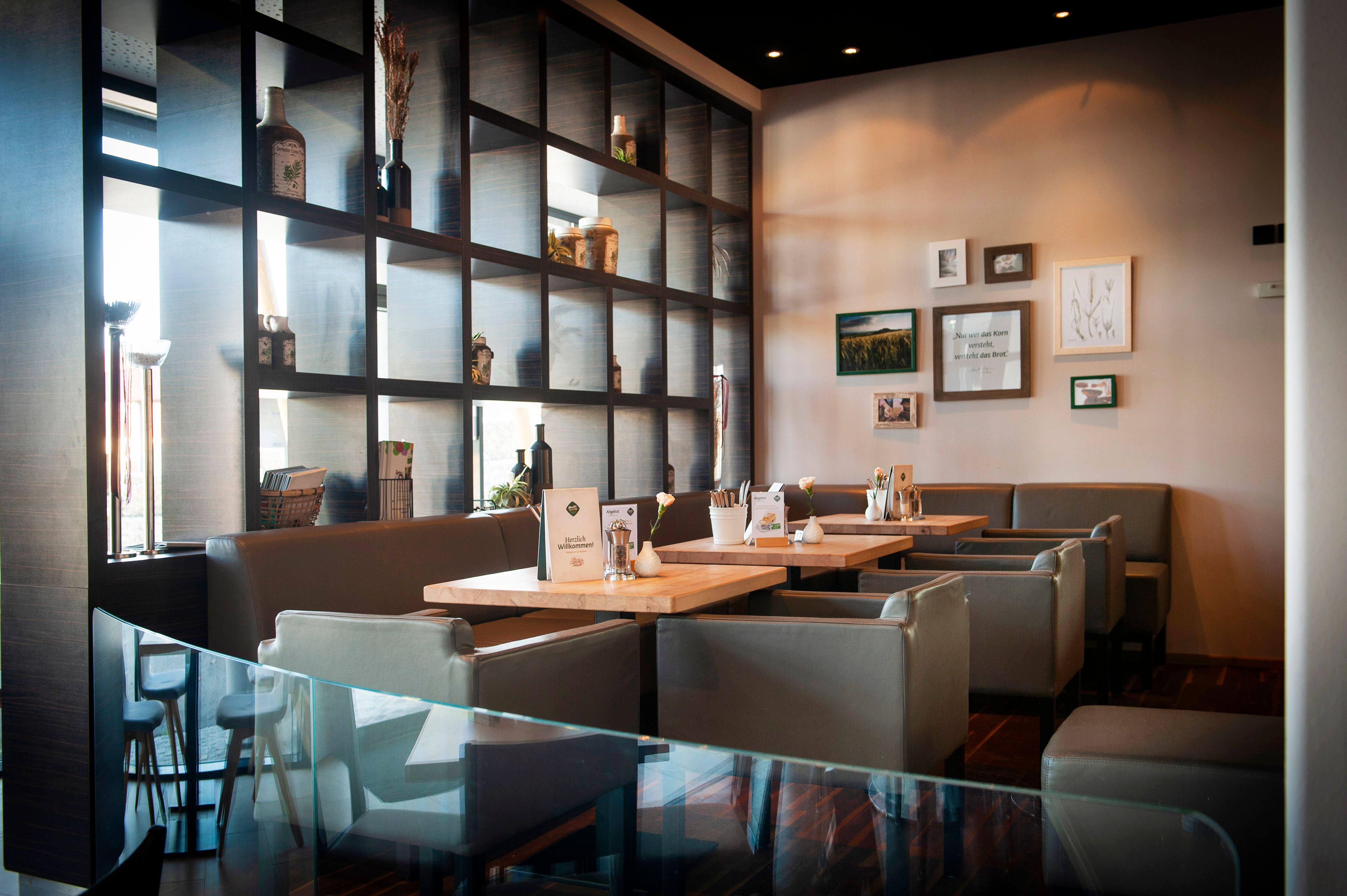 Innenansicht eines modernen Cafés mit Holztischen und grauen Stühlen, dekoriert mit Pflanzen und Bildern an der Wand.