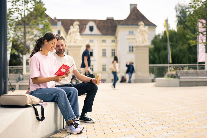 Ein Paar sitzt auf einer Bank im Freien und liest gemeinsam ein Buch. Im Hintergrund ist ein historisches Gebäude zu sehen.