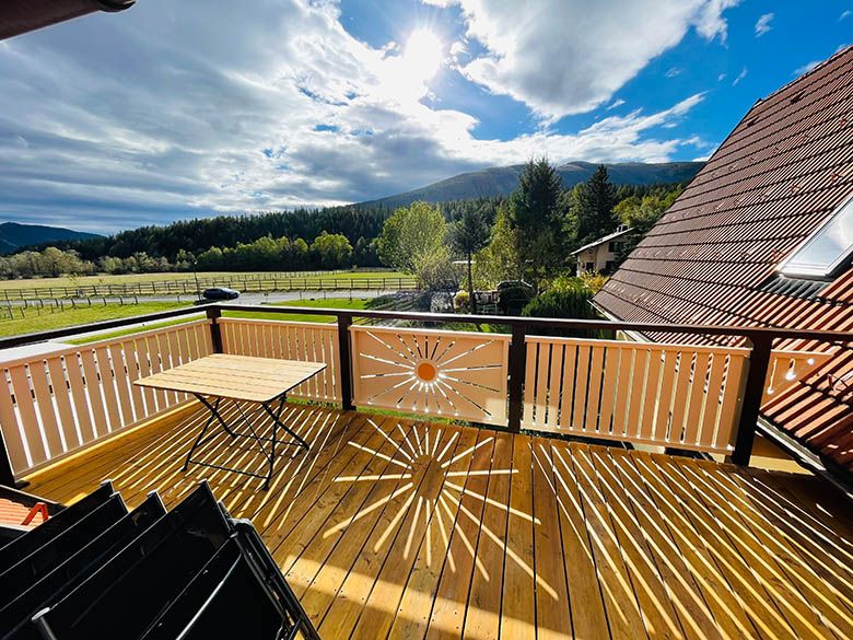 Holzbalkon mit Tisch, Blick auf Wiesen und Berge, Sonnenstrahlenmuster auf dem Boden.