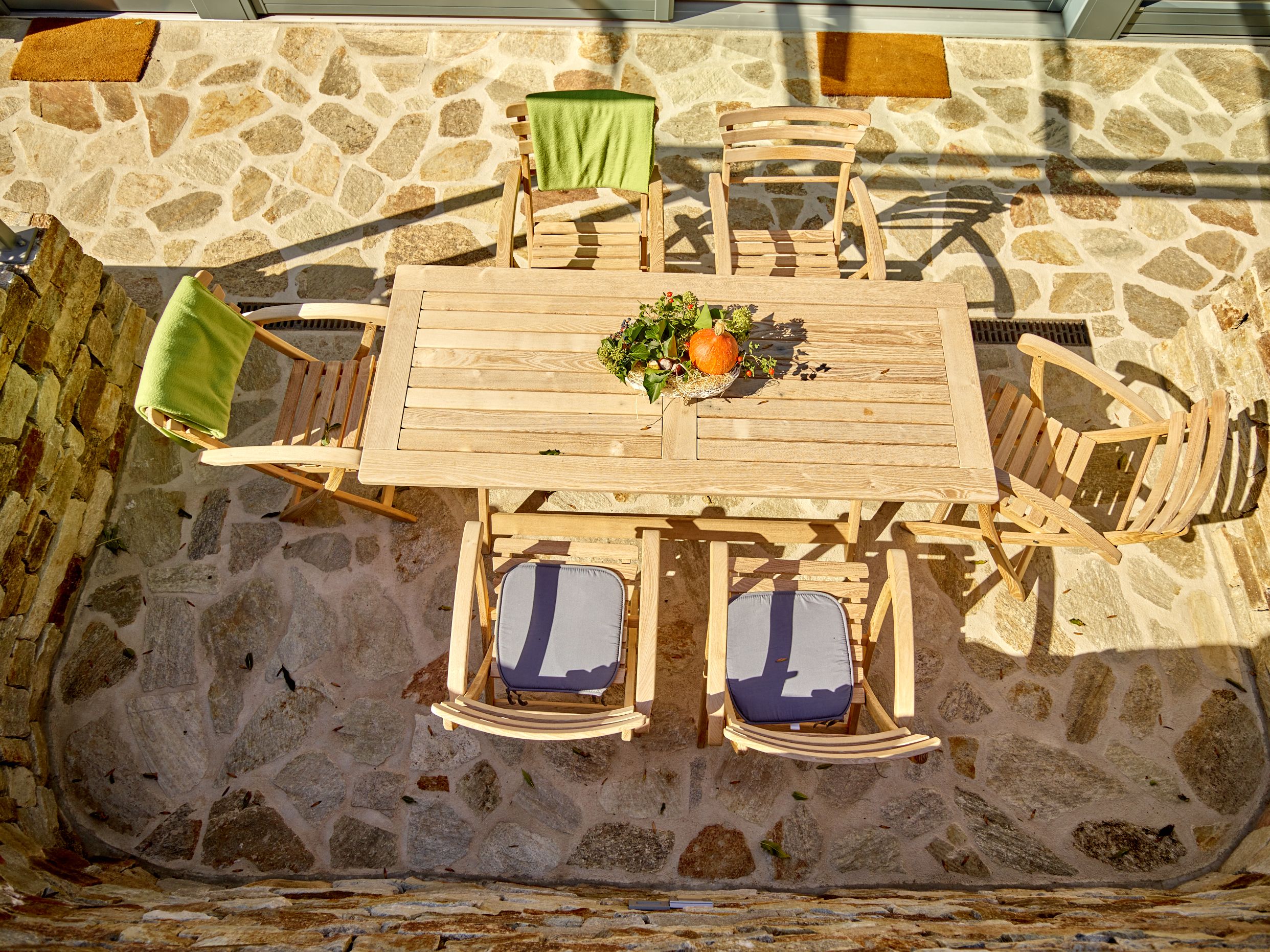 Holztisch mit Stühlen auf einer Steinterrasse, dekoriert mit Kürbis und Blumen.