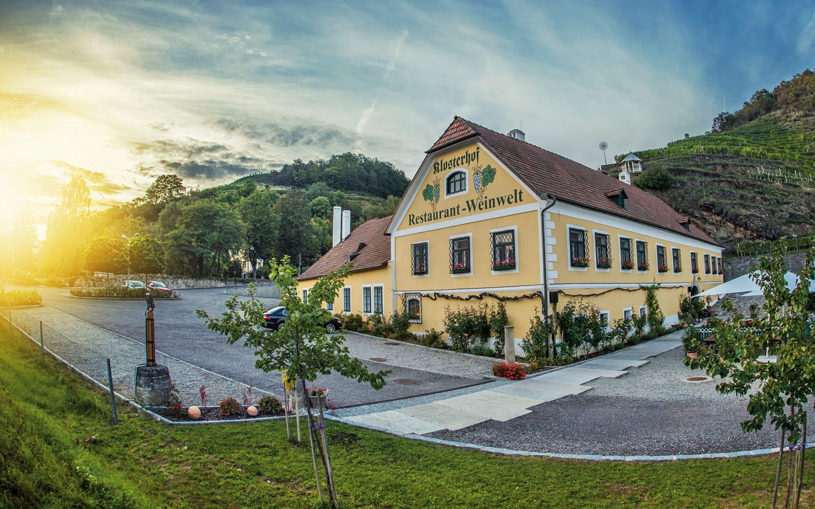 Außenansicht eines gelben Gebäudes mit der Aufschrift 'Klosterhof Restaurant-Weinwelt', umgeben von Bäumen und Weinbergen bei Sonnenuntergang.
