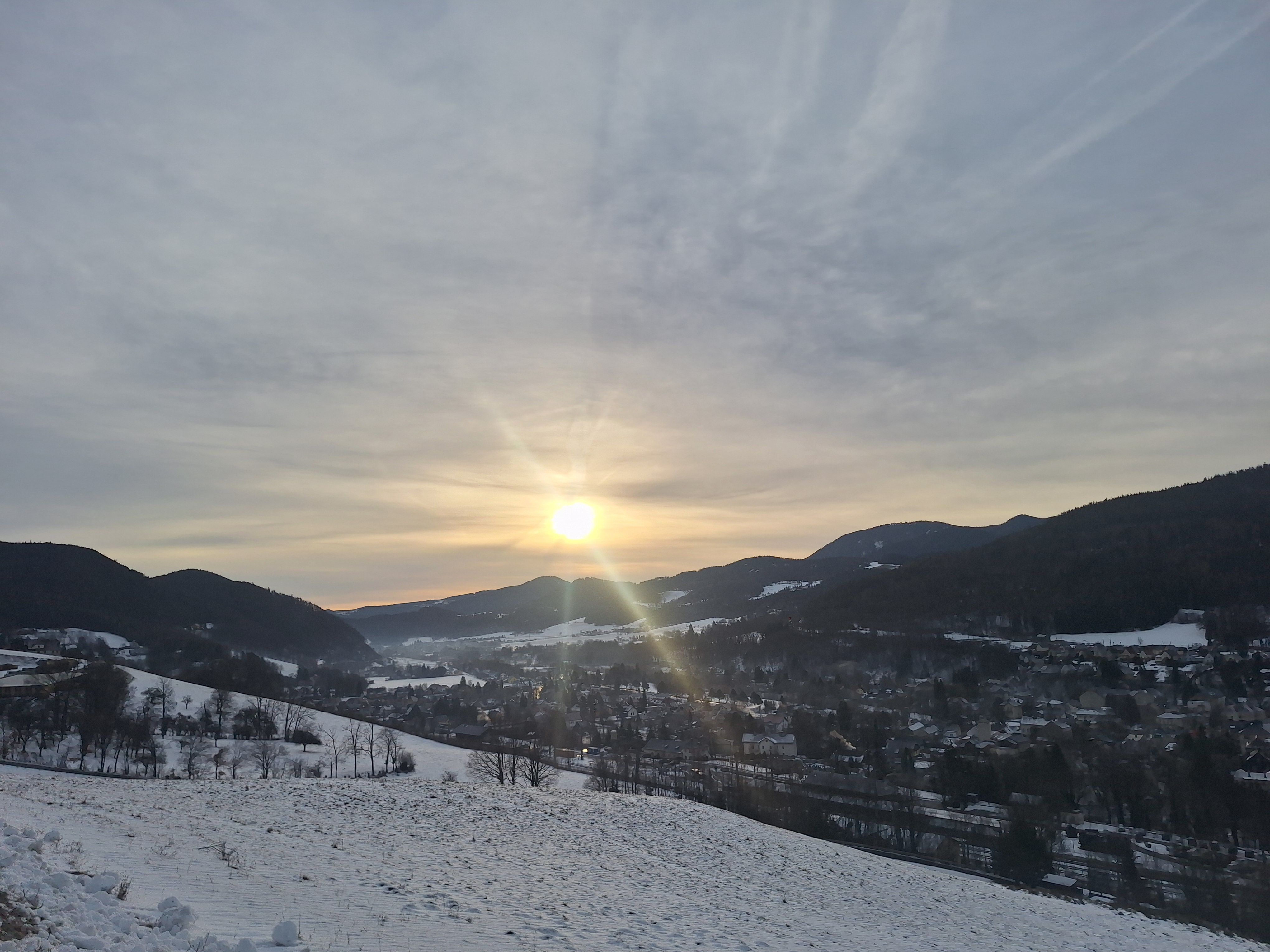 Winterlandschaft in Payerbach mit Sonnenaufgang über schneebedeckten Hügeln und einem Dorf im Tal.