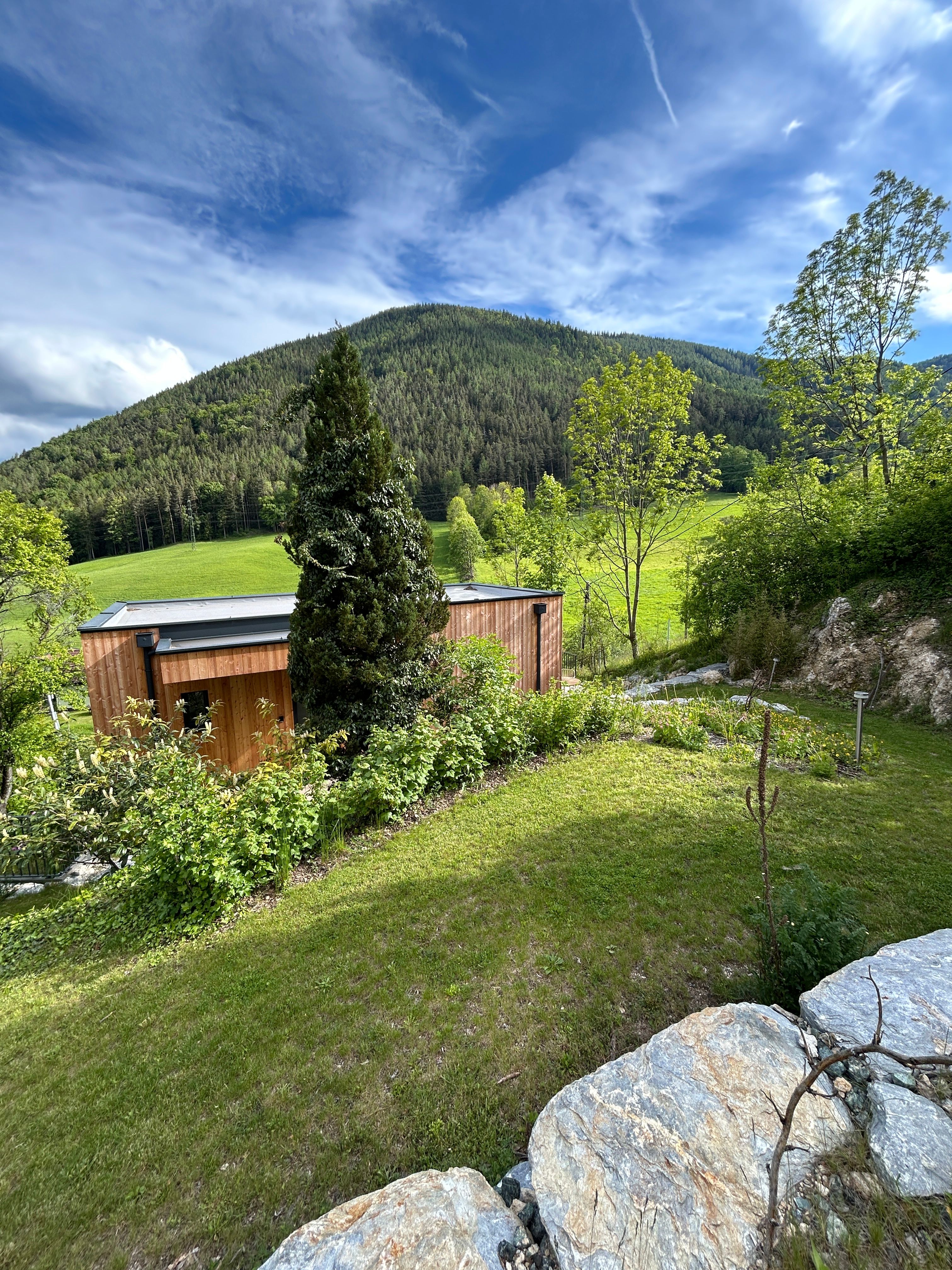 Ein kleines Holzhaus steht in einer grünen Landschaft mit einem bewaldeten Hügel im Hintergrund und einem blauen Himmel darüber.