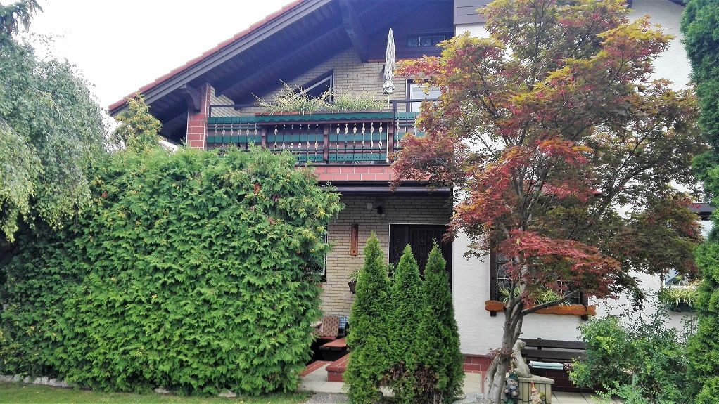 Außenansicht eines Hauses mit Balkon, umgeben von Bäumen und Sträuchern.