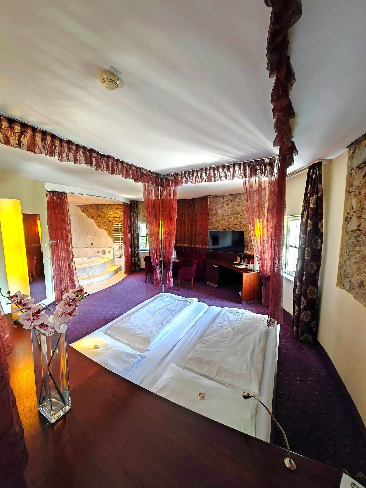 Ein luxuriöses Hotelzimmer mit einem großen Bett, roten Vorhängen, einem Fernseher und einer Badewanne im Hintergrund.