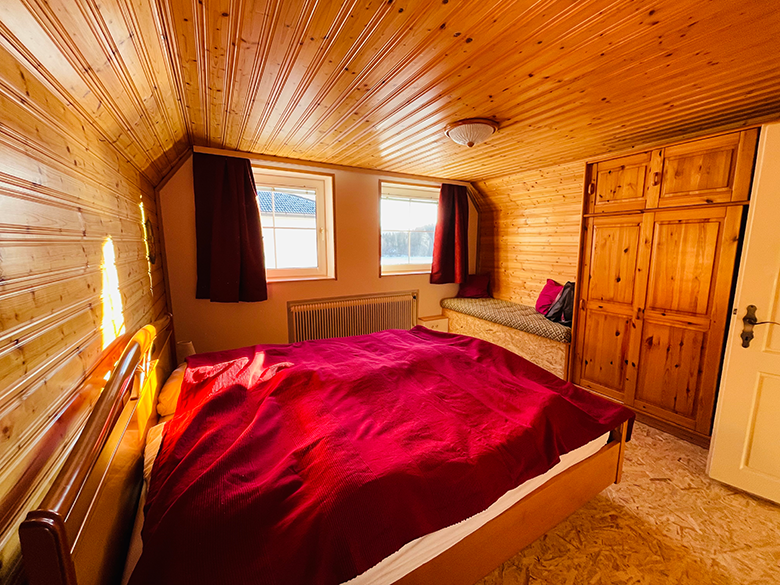 Holzvertäfeltes Schlafzimmer mit rotem Bettbezug, zwei Fenstern, einem Schrank und einer Sitzbank.