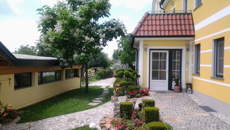 Eingang eines gelben Geb&auml;udes mit gepflegtem Garten und gepflastertem Weg.