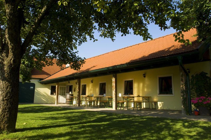 Ein Heurigengarten mit gelbem Gebäude, roten Dachziegeln und grüner Wiese.