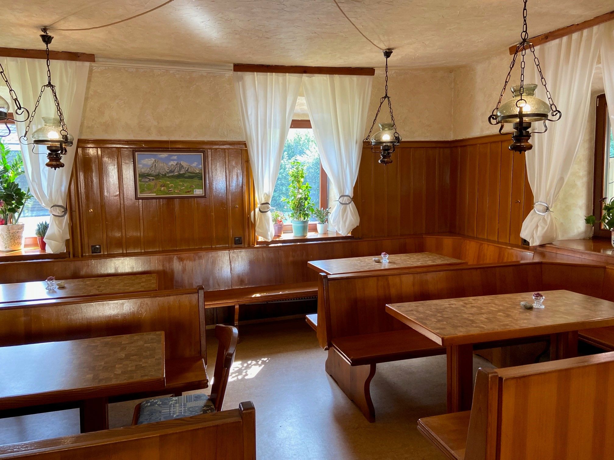 Gemütliche Gaststube mit Holzmöbeln, weißen Vorhängen und Pflanzen auf der Fensterbank.