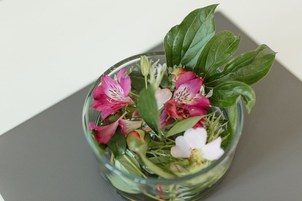 Ein Glasgefäß mit bunten Blumen und grünen Blättern auf einem Tisch.