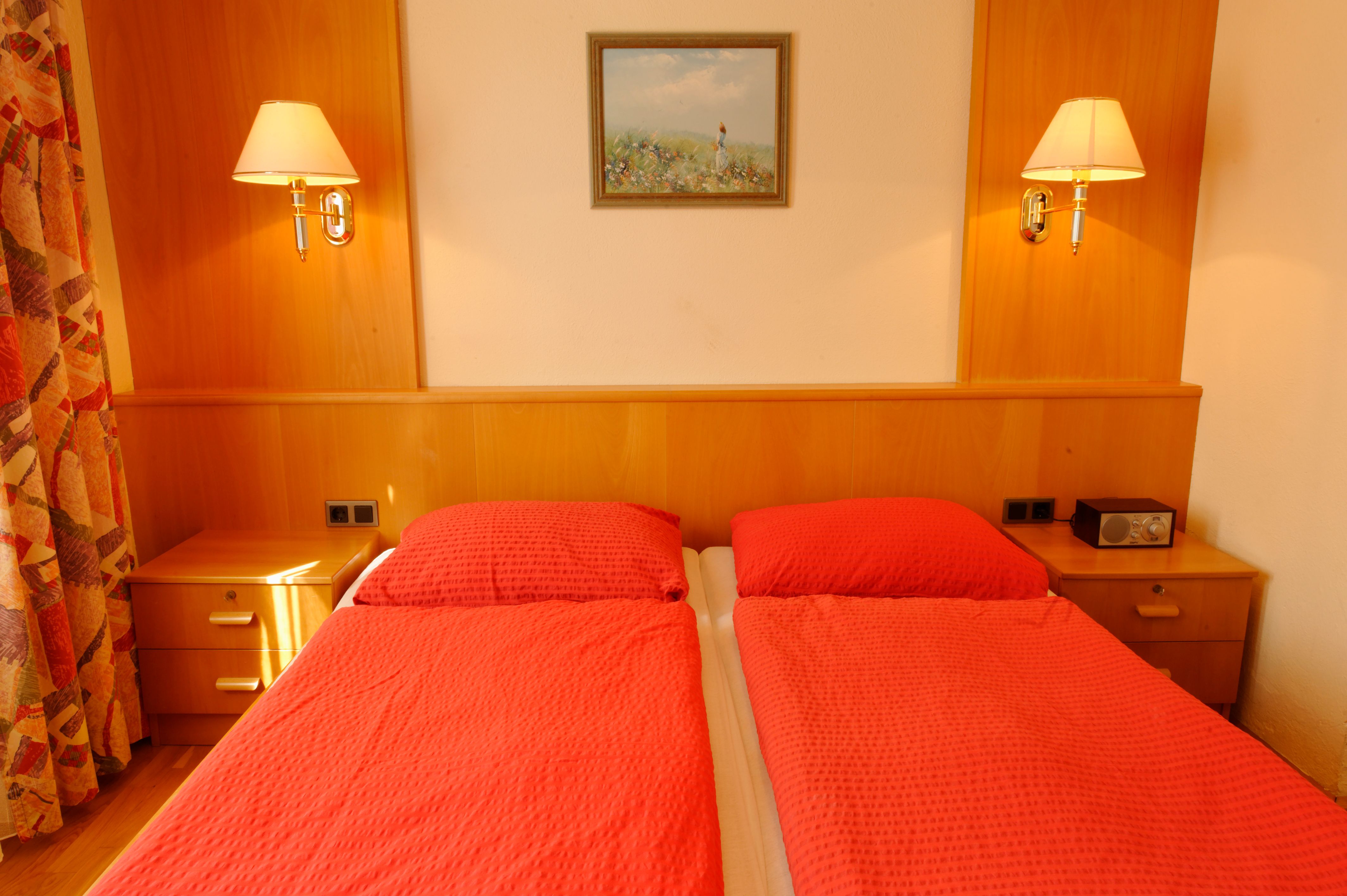 Gästezimmer mit zwei Einzelbetten, roten Bettdecken, Holzmöbeln und Wandlampen.