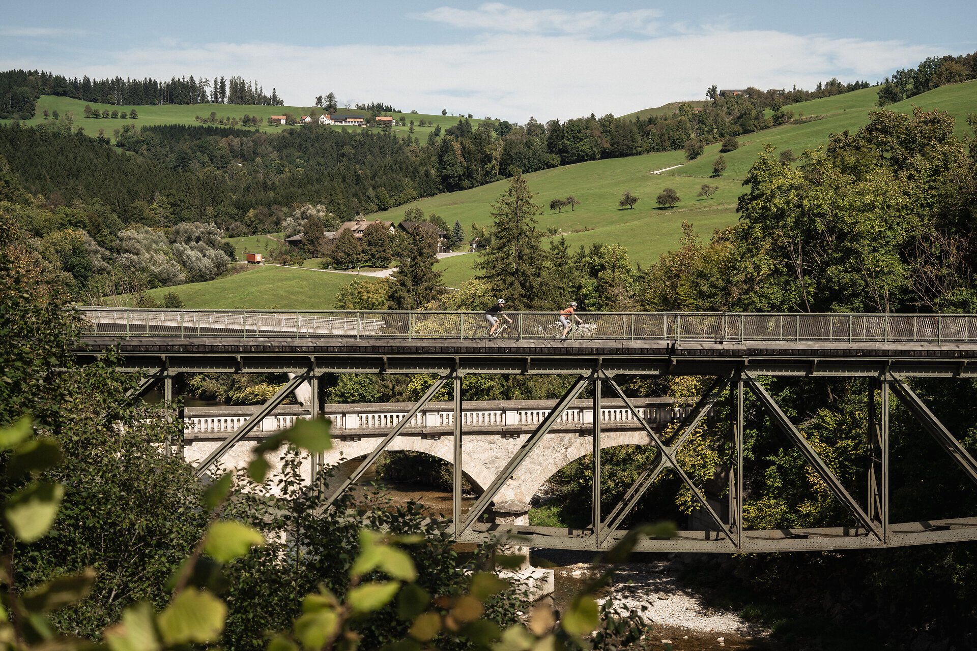 Das Bild zeigt eine idyllische Landschaft entlang des Ybbstalradwegs, in der zwei Radfahrer:innen über eine Brücke fahren, umgeben von grünen Wiesen, Bäumen und einem klaren Himmel, der ein Gefühl von Freiheit und Naturverbundenheit vermittelt.