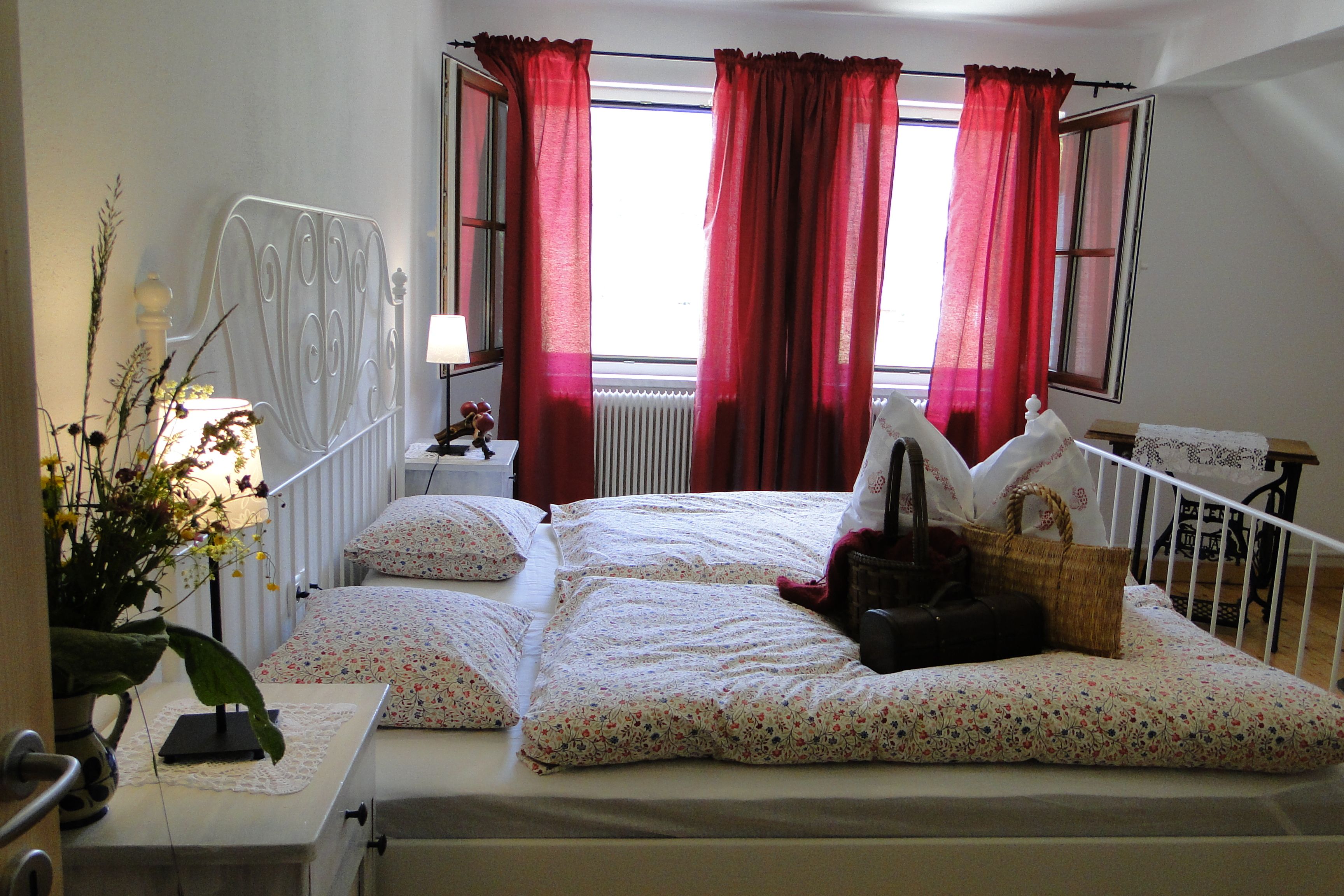 Gemütliches Schlafzimmer mit Doppelbett, roten Vorhängen und Blumen auf dem Nachttisch.