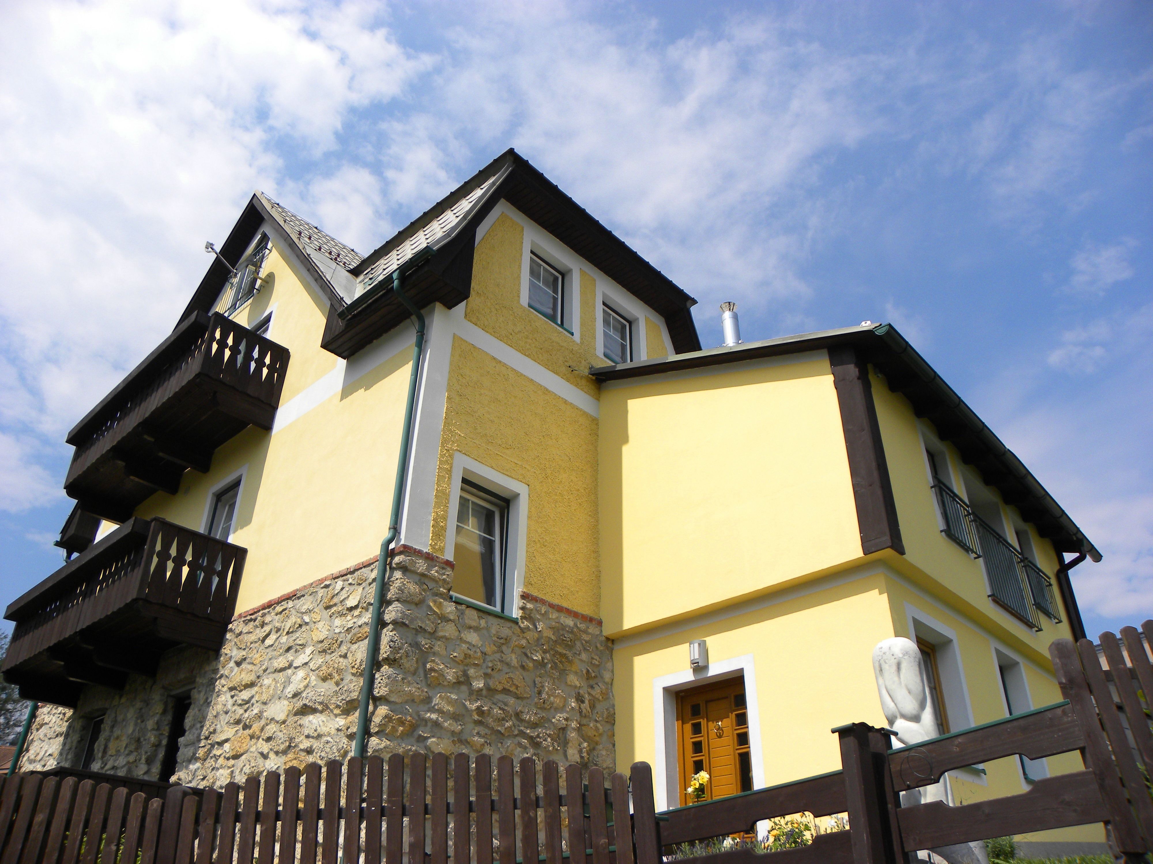 Gelbes Haus mit Holzdetails und Steinmauer, blauer Himmel im Hintergrund.
