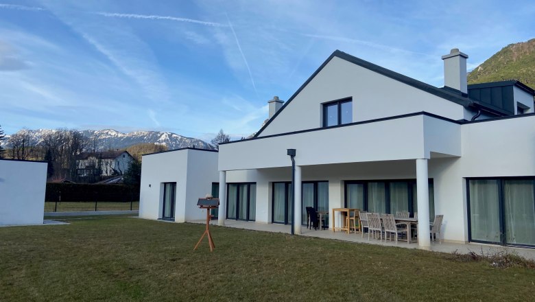 Modernes wei&szlig;es Haus mit Terrasse und Garten, im Hintergrund schneebedeckte Berge.