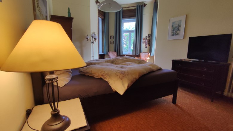 Gemütliches Schlafzimmer mit Doppelbett, Nachttischlampe, Fenster und Fernseher.