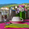 Fr&uuml;hst&uuml;ckstisch im Freien mit rosa Tischsets, Kaffeetassen, Salz- und Pfefferstreuern und Blumenarrangement, im Hintergrund eine malerische Landschaft.