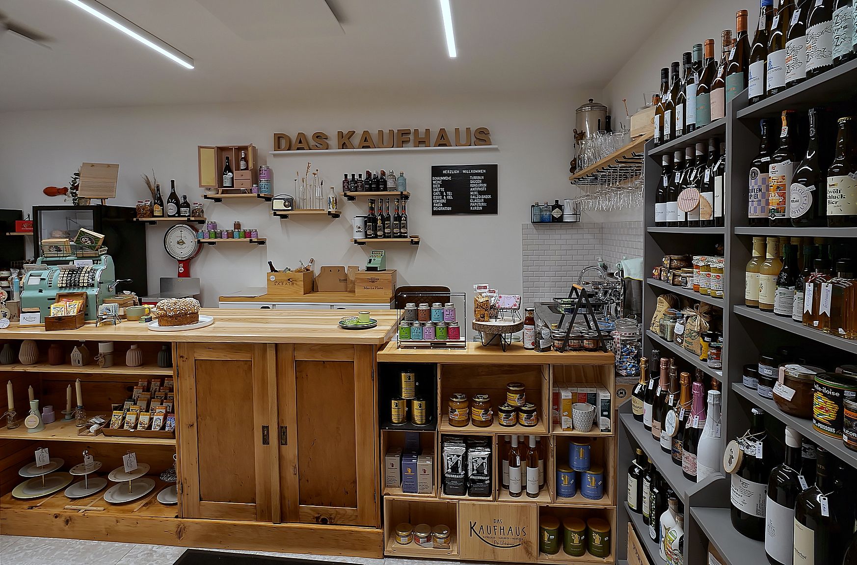 Innenansicht eines kleinen, gemütlichen Ladens mit Holzregalen, gefüllt mit Weinflaschen, Lebensmitteln und einer alten Registrierkasse.