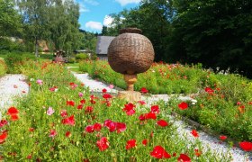 Ein Garten mit roten und rosa Mohnblumen, einem gro&szlig;en, runden Korb auf einem Holzsockel in der Mitte und B&auml;umen im Hintergrund.