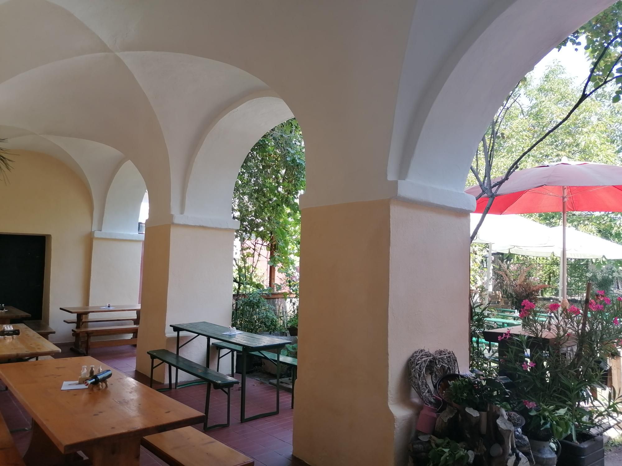 Arkaden mit Holzbänken und Tischen, Pflanzen und Sonnenschirmen im Hintergrund.