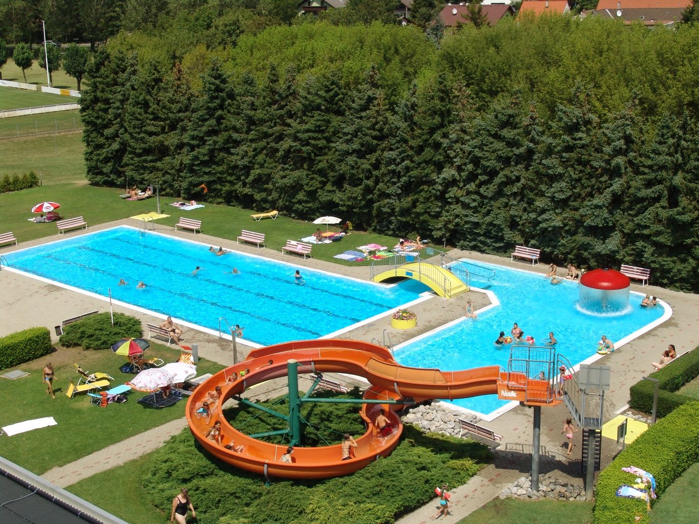 Luftaufnahme eines Freibads mit Rutsche, zwei Pools und Liegewiese.
