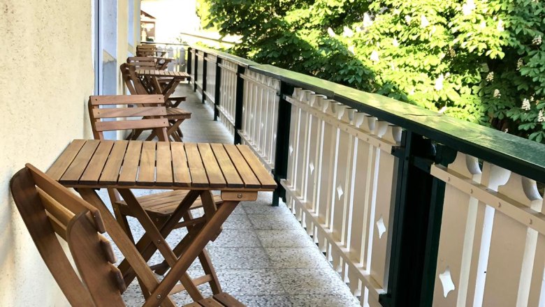 Holzbalkon mit Tisch und Stühlen, daneben grüne Bäume.