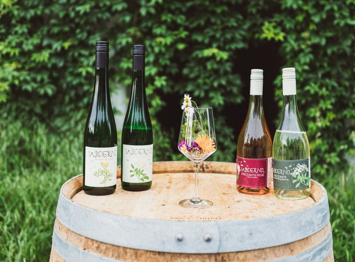 Vier Weinflaschen und ein Weinglas mit Blumen auf einem Holzfass vor grüner Hecke.