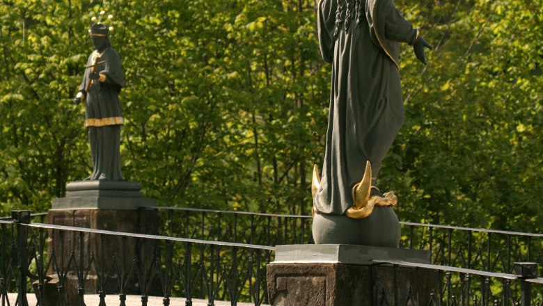 Zwei Statuen auf einer Brücke mit Geländer, umgeben von grünen Bäumen.