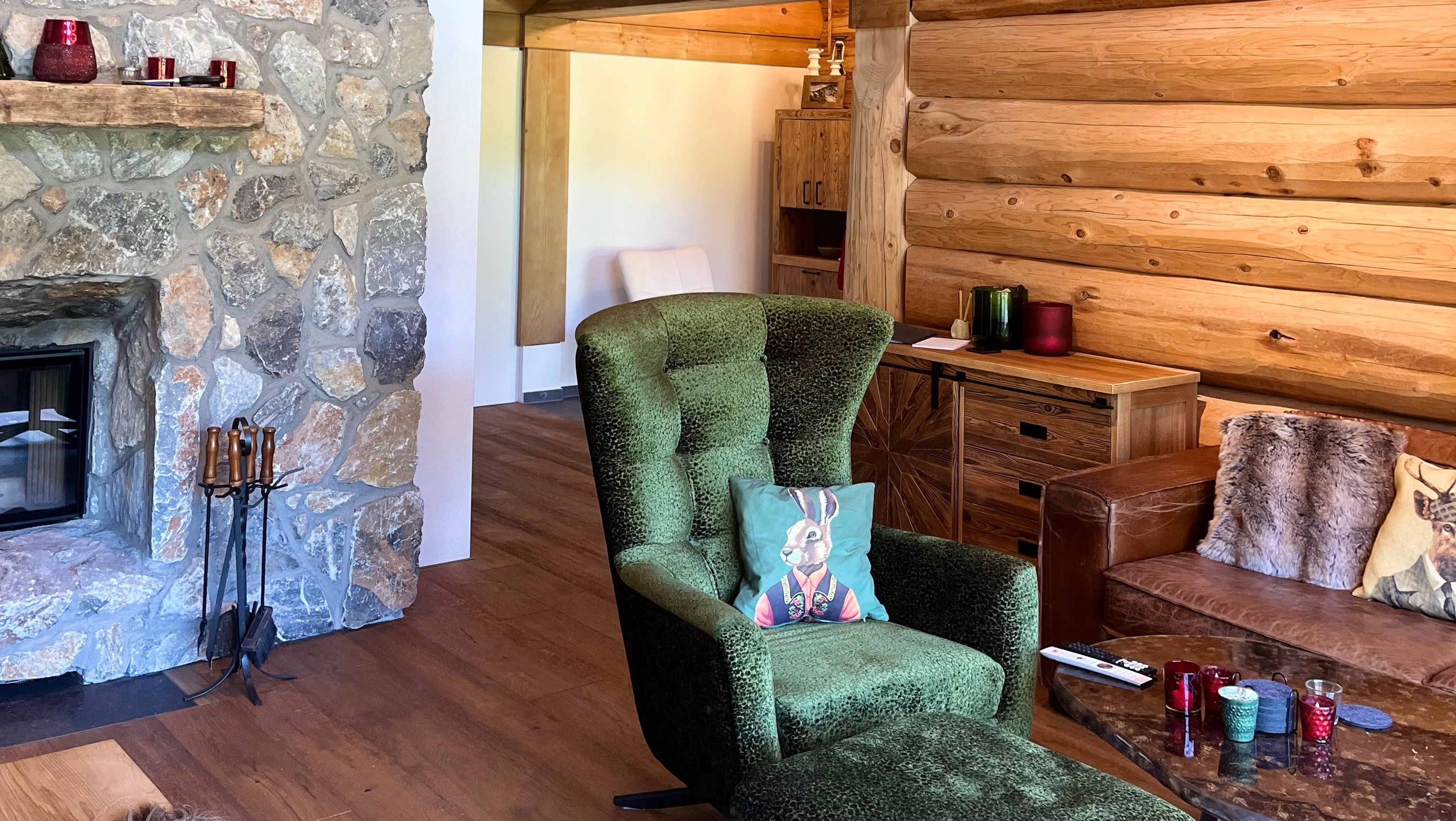 Gemütliches Wohnzimmer mit Steinwand und Kamin, grünem Sessel, Holzmöbeln und dekorativen Kissen.