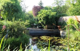 Ein idyllischer Garten mit Teich, Seerosen und üppiger Vegetation. Im Hintergrund stehen ein Tisch und Stühle auf einer Terrasse.