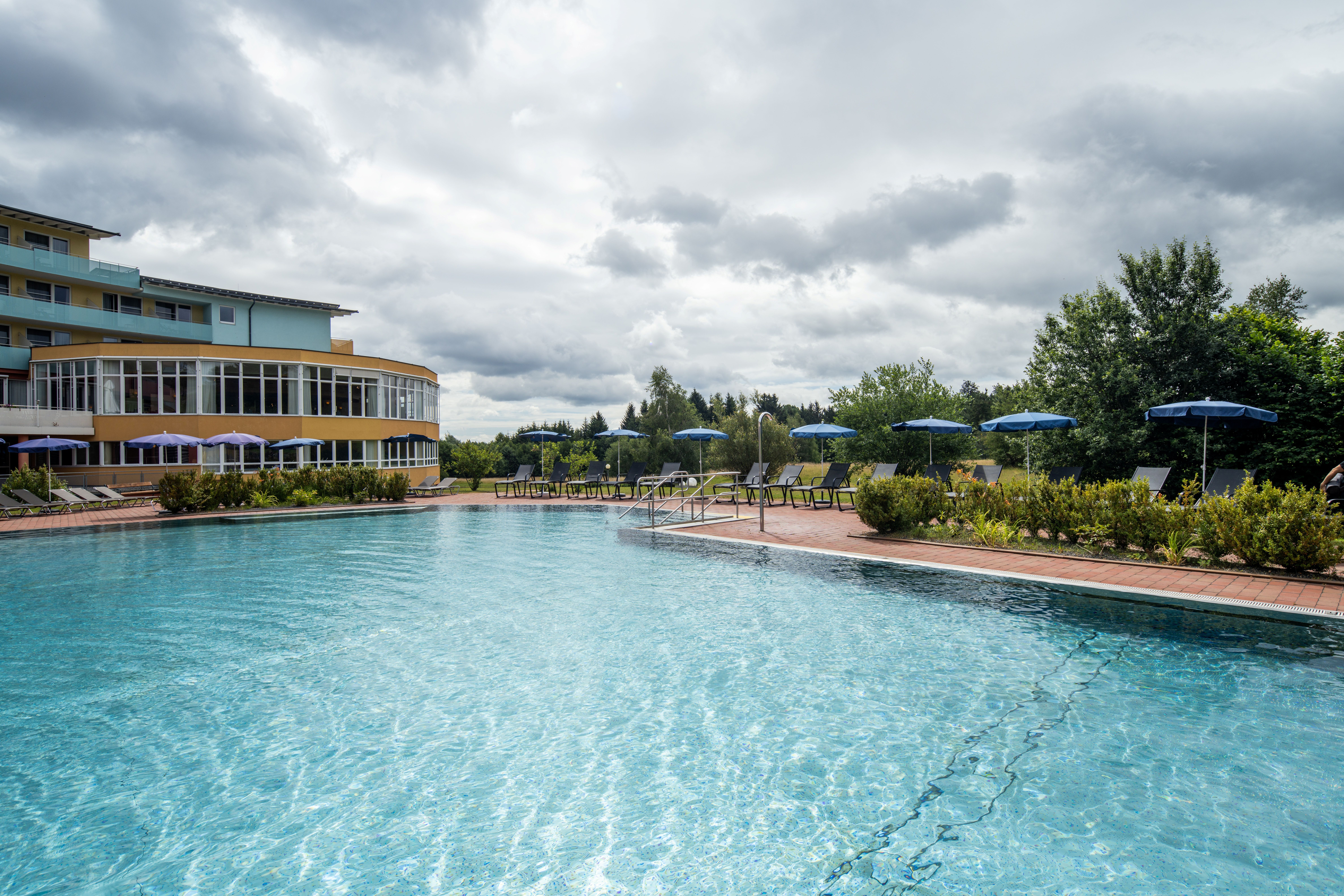 Ein Außenpool mit klarem Wasser vor einem Gebäude mit großen Fenstern und mehreren Stockwerken. Am Pool stehen Liegestühle und Sonnenschirme.