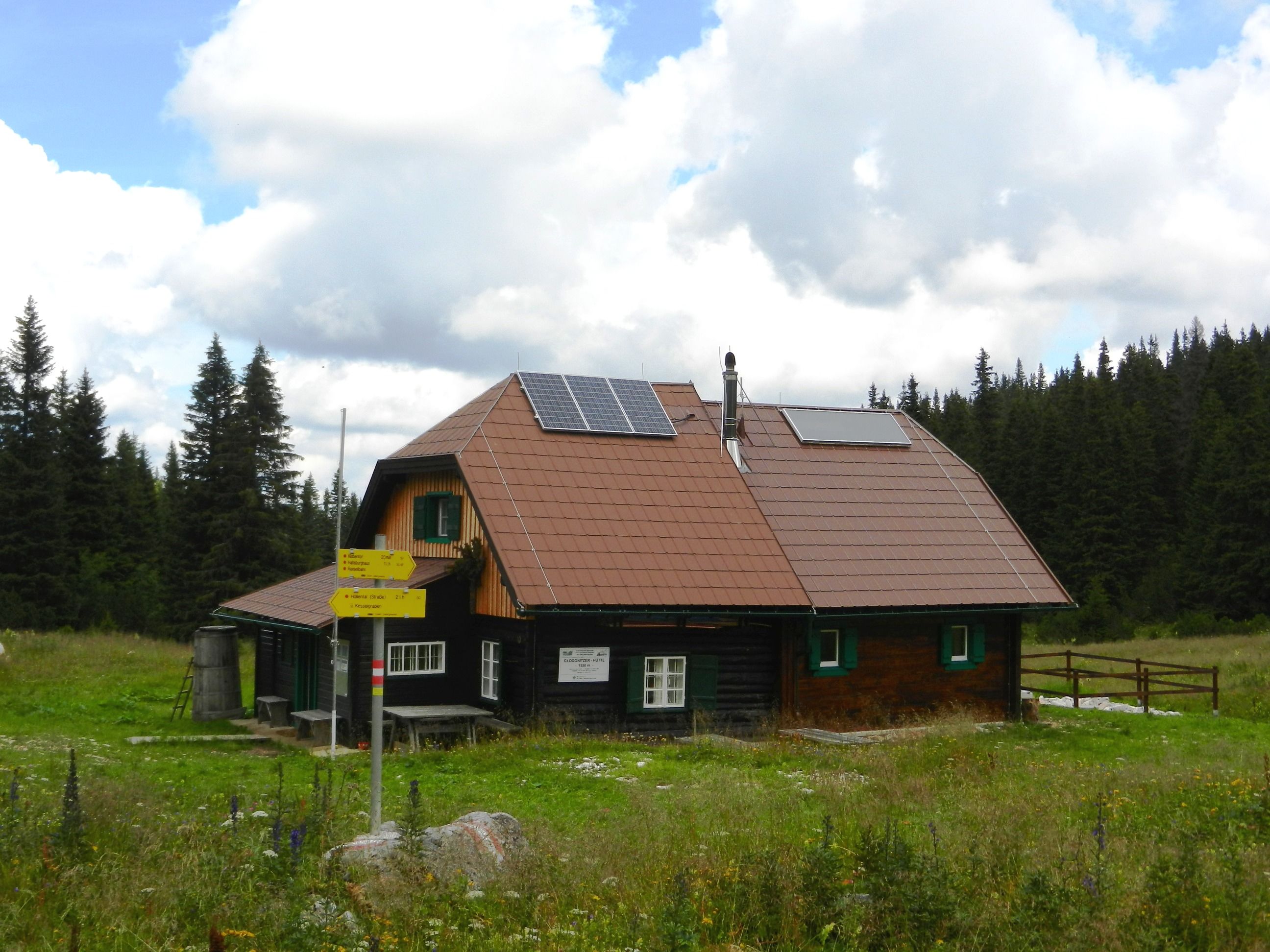 Eine Berghütte mit Solarpanelen auf dem Dach, umgeben von Wiesen und Wald.