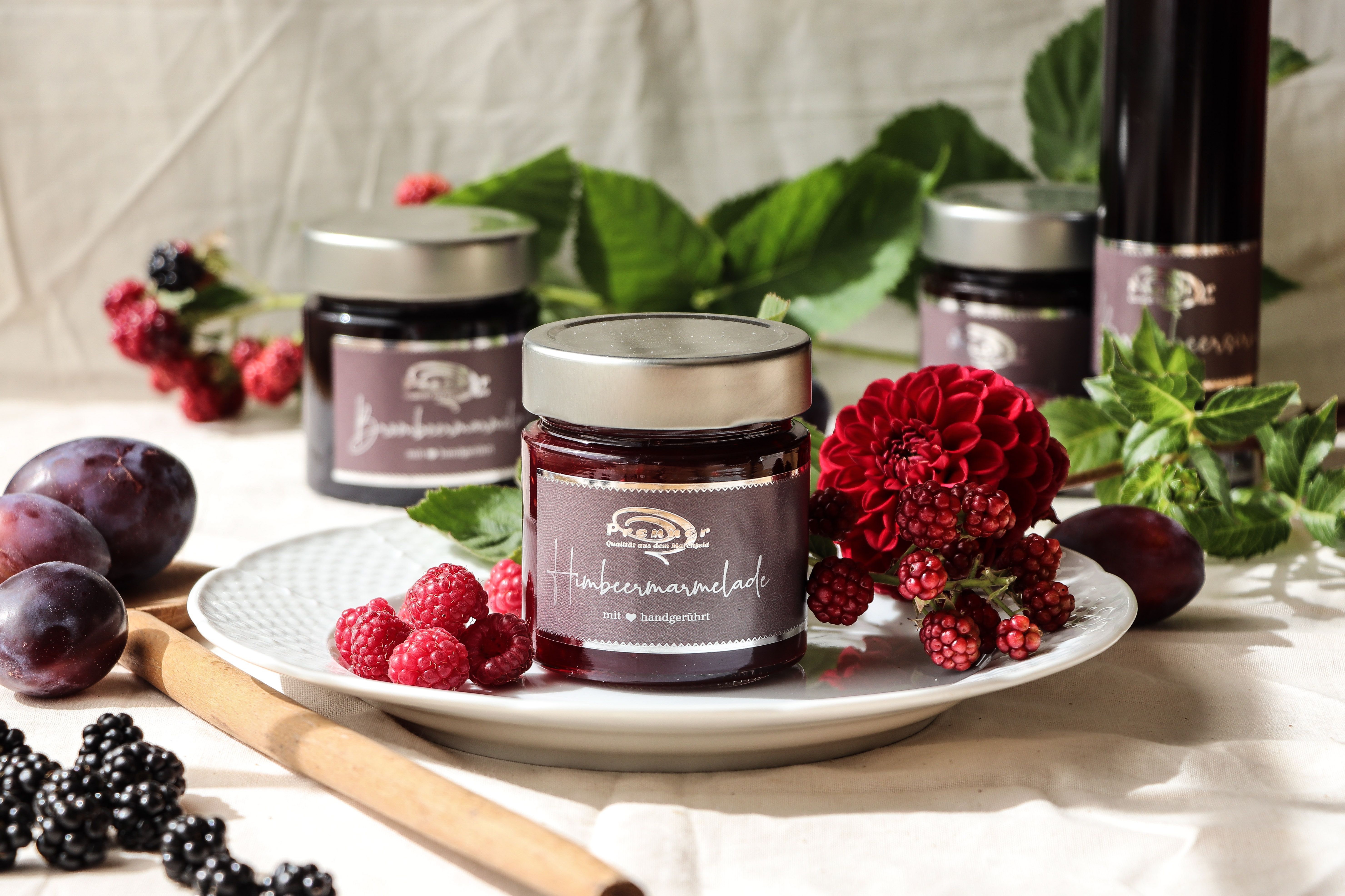 Ein Glas Himbeermarmelade auf einem Teller mit frischen Beeren und Blumen dekoriert.