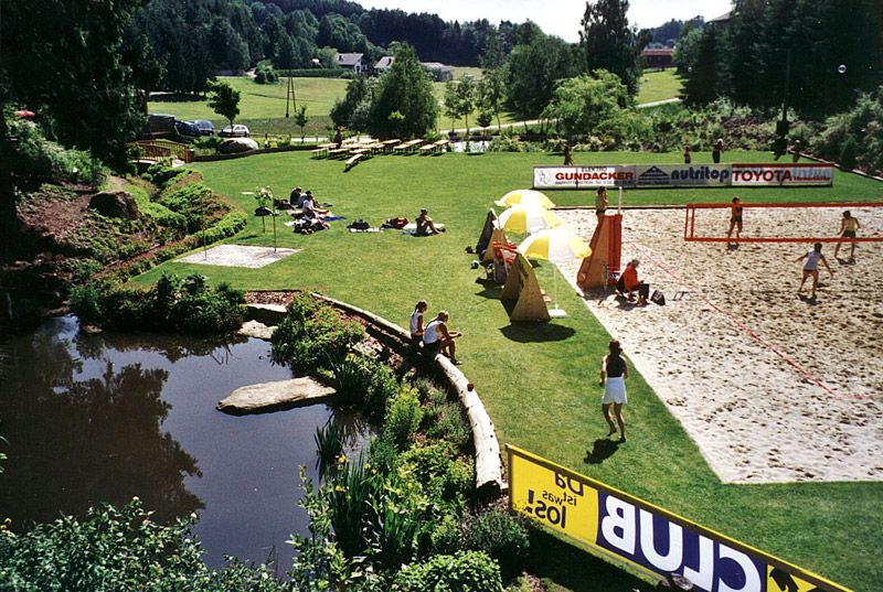 Grüne Wiese mit Badeteich, Volleyballfeld und Menschen beim Entspannen und Spielen.