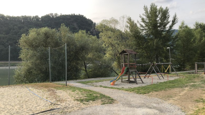 Spielplatz mit Rutsche und Schaukeln, umgeben von B&auml;umen und H&uuml;geln.